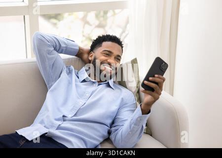 Young handsome smiling man lying on sofa with smartphone Stock Photo
