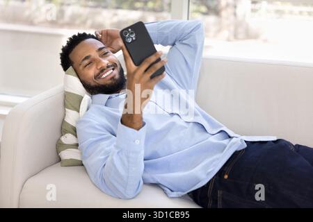 Relaxed African guy using smartphone while lying on cozy sofa Stock Photo