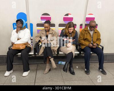 People sit on a bench using smartphones at a public transport station, reflecting digital habits during everyday urban commuting. Stock Photo