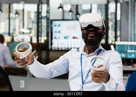 Male corporate employee using VR headset and wireless controllers, interacting with 3D virtual data and numbers. Black man working in modern high tech IT company with futuristic digital technologies. Stock Photo
