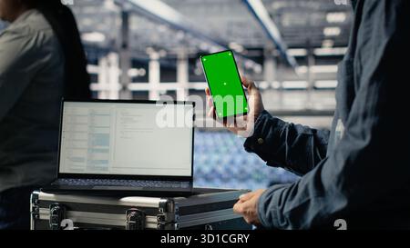 Server room software developer using mockup phone while waiting for updates to finish installing. Data center engineer uses green screen smartphone, waiting for code to finish compiling, camera B Stock Photo