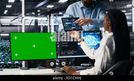Data center manager overseeing team of technicians managing AI systems using green screen PC. Supervisor checking work of teamworking server hub colleagues using mockup computer, camera A Stock Photo