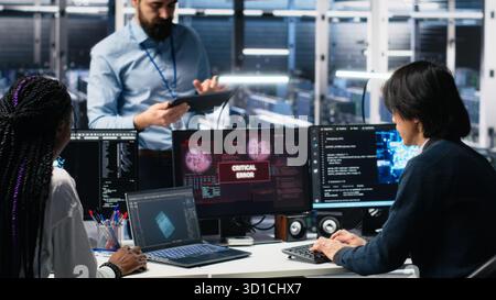 Data center manager coordinating programmers fixing critical error while using AI. Server room team leader overseeing workers repairing glitch popup while using artificial intelligence, camera A Stock Photo