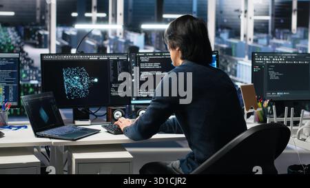 Data center technician entering office, managing AI systems for uptime and efficiency. IT professional arriving at work, sitting at desk, using artificial intelligence in server hub, camera A Stock Photo
