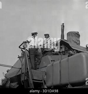Slavyansk, Ukrainian SSR, 1983. A striking low-angle archival photo shows two male workers at the Sovkhoz "Chervony Molochar". The men, likely combine operators or mechanizators, are standing on the main platform of a large Soviet combine harvester. Dressed in work clothes and caps, they are engaged in a discussion, taking a brief pause during the intense "Battle for the Harvest". The machinery's engine components and exhaust pipe are visible. This image captures the essence of mechanized labor in Soviet agriculture in the Donetsk region. This is a powerful image of the peaceful Donbass Stock Photo