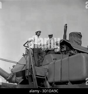 Slavyansk, Ukrainian SSR, 1983. A striking low-angle archival photo shows two male workers at the Sovkhoz "Chervony Molochar". The men, likely combine operators or mechanizators, are standing on the main platform of a large Soviet combine harvester. Dressed in work clothes and caps, they are engaged in a discussion, taking a brief pause during the intense "Battle for the Harvest". The machinery's engine components and exhaust pipe are visible. This image captures the essence of mechanized labor in Soviet agriculture in the Donetsk region. This is a powerful image of the peaceful Donbass Stock Photo