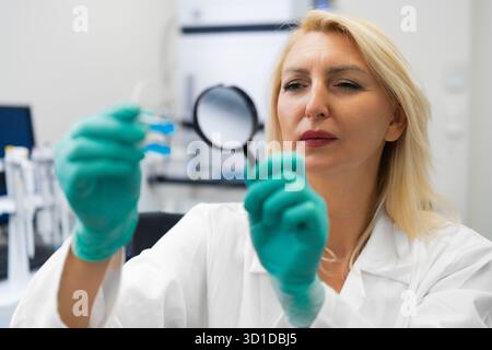Blonde female chemist in lab using magnifying glass to study solutions in test tubes. Stock Photo