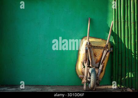 Old yellow rusty wheelbarrow propped up against a vibrant green wall Stock Photo