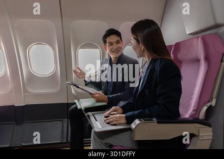 A business man and woman in formal suits working together on a flight. They are discussing documents and using laptops, showing teamwork and productiv Stock Photo