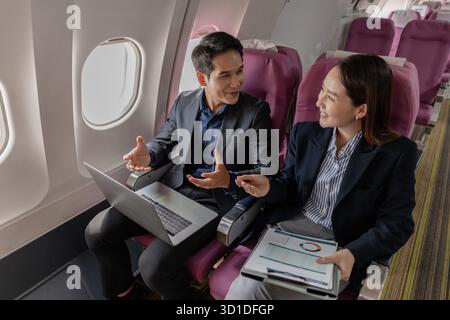 A business man and woman in formal suits working together on a flight. They are discussing documents and using laptops, showing teamwork and productiv Stock Photo