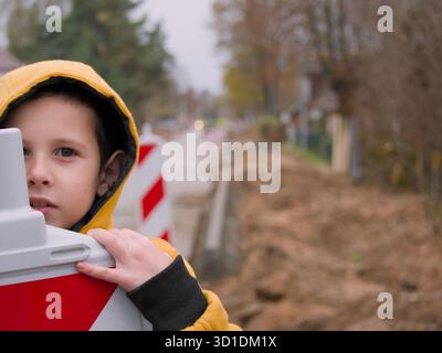 Child Peeking from Barrier - A curious child plays at a road work site, looking from behind a safety barrier - Kid at construction site Stock Photo