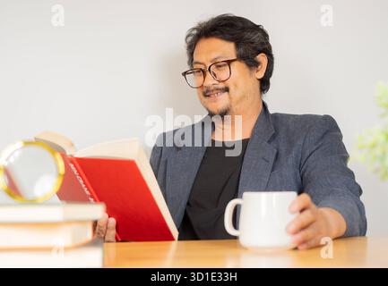 Handsome man with a beard and coffee reading a book sitting at desk in the living room. Education and success concept. Stock Photo