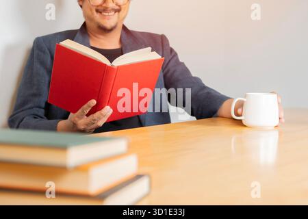 Happy Asian businessman in casual suit, holding coffee cup and reading a book while sitting in reading room. Selective focus. Stock Photo