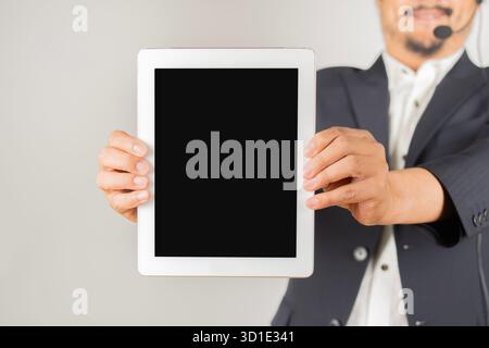 Adult Asian business man call center wearing headset showing a tablet with blank screen standing on white background, agent with support and service o Stock Photo