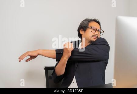 Man stretches during work break. Adult Asina man in suit sitting by desk with computer in home office and doing relaxing exercise to prevent muscle pa Stock Photo