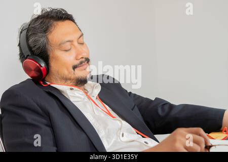 Businessman resting inside office with headphones listening to music and audio books podcasts with eyes closed dozing. Stock Photo