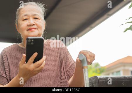 Portrait image of cheerful senior woman sitting at outdoor and holding walking stick while using her smart phone. Retirement Healthy Concept. Stock Photo