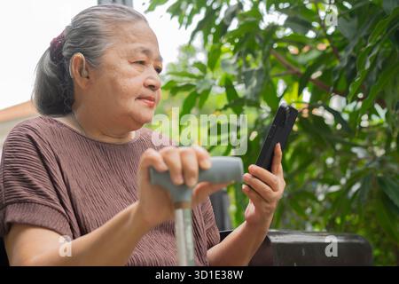Happy retired woman with using mobile phone and walking stick in the garden at home. Age care concept. Stock Photo