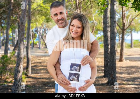 Pregnant couple hugging and holding ultrasound photo outdoors in green park Stock Photo