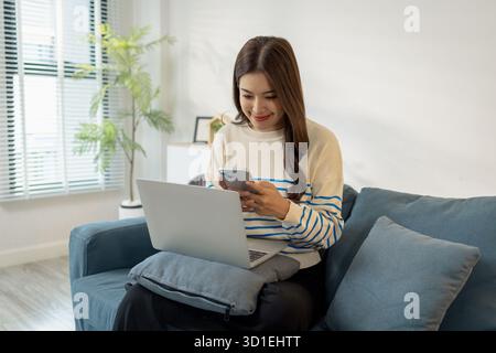 A young woman sits comfortably on the sofa, smiling while using her smartphone with a laptop in front of her, representing digital lifestyle, connecti Stock Photo