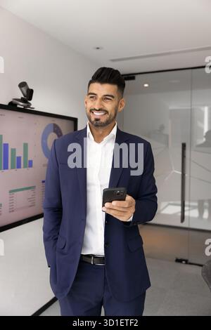 Happy dreamy young Indian marketing expert man holding smartphone Stock Photo
