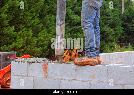 Worker stands on wall, directing concrete into block structure using pipe while surrounded by equipment. Stock Photo
