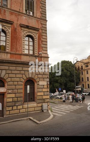 Corner of a classic Roman building with red and tan stone, looking across a zebra crossing and a group of pedestrians on a street corner. Stock Photo