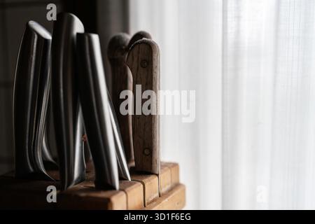Stainless steel kitchen knives and a wooden handle knife standing in a wooden knife block, receiving soft natural light from a window covered by a she Stock Photo