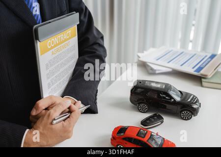 A businessman in a suit is calculating loan details, reviewing financial documents, and holding car keys near a calculator and toy car models, represe Stock Photo