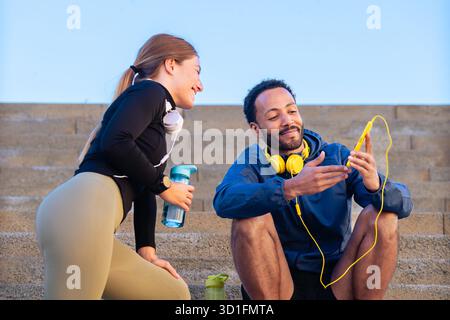 Two friends resting on stairs after workout, smiling, sharing music and water. Stock Photo