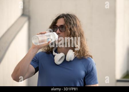 Handsome sporty fit athlete drinking water from bottle in sunny day against urban white wall. Water balance. Stock Photo
