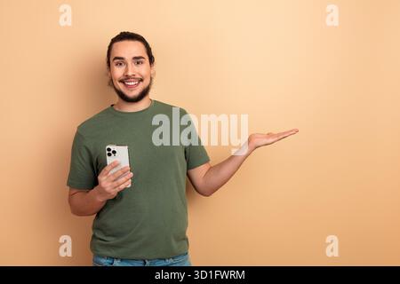 Young man with green tshirt using smartphone smiling beside beige backdrop Stock Photo