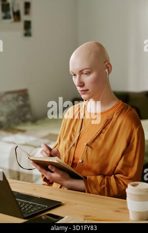 Young Adult Woman with Alopecia Writing in Notebook While Listening Stock Photo