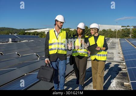 Team of diverse energy engineers and technicians inspecting solar panels on a rooftop, discussing clean energy solutions and future renewable power ge Stock Photo