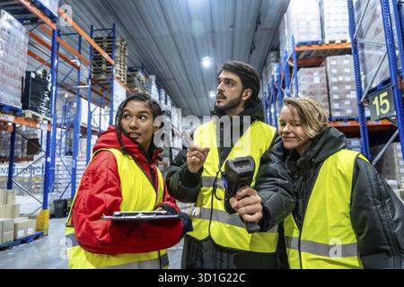 Diverse team of warehouse workers wearing safety vests and warm clothing, using a barcode scanner and clipboard while inspecting products in a cold st Stock Photo