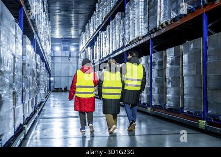 Three workers wearing safety vests and warm clothing are inspecting goods in a modern industrial cold storage warehouse, ensuring proper inventory and Stock Photo