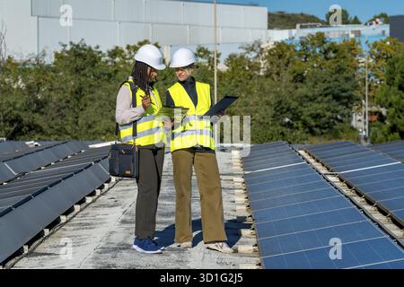 Two female engineers wearing hard hats and reflective vests are reviewing documents while standing on a rooftop next to solar panels, highlighting ren Stock Photo