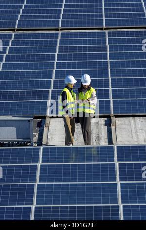 Female engineers wearing hard hats and high visibility vests discussing data on a digital tablet while standing on a rooftop solar panel installation Stock Photo