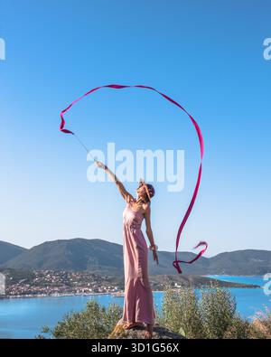 Beautiful woman in silk rosa dress standing on a scenic viewpoint, gracefully performing with a ribbon like a rhythmic gymnast during a summer holiday Stock Photo