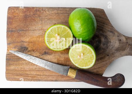 Close-up of fresh green limes (whole and sliced) on a rustic wooden board with a knife. White background provides copy space. Cooking concept. Stock Photo