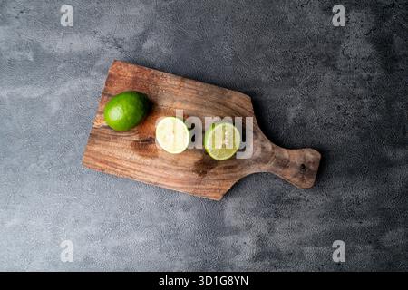 Close-up of fresh green limes, whole and sliced. The citrus fruit is on a rustic wooden cutting board with a dark, textured background. Stock Photo