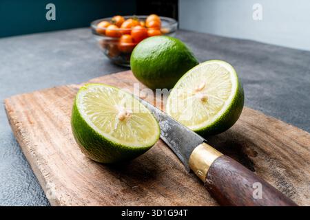 Close-up of fresh green limes, whole and sliced. The citrus fruit is on a rustic wooden cutting board with a dark, textured background. Stock Photo