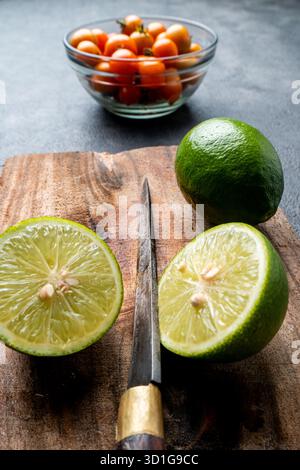 Close-up of fresh green limes, whole and sliced. The citrus fruit is on a rustic wooden cutting board with a dark, textured background. Stock Photo