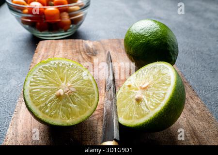 Close-up of fresh green limes, whole and sliced. The citrus fruit is on a rustic wooden cutting board with a dark, textured background. Stock Photo