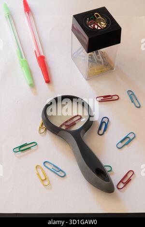Magnifying glass, colorful paper clips, and pens arranged on white paper background Stock Photo