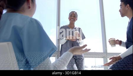 Chatting medical team in scrubs holding two paper coffee cups by floor-to-ceiling windows in lounge Stock Photo