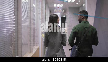 Walking two professionals side-by-side down office hallway, with notebook, messenger bag, blinds Stock Photo