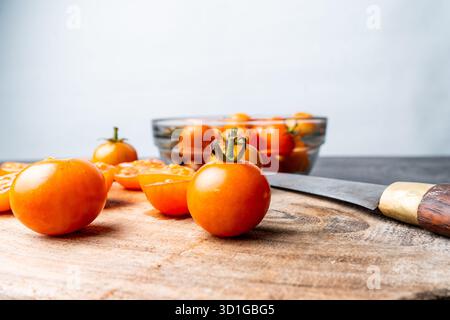 Close-up of fresh cherry tomatoes, whole and sliced, on a rustic wooden cutting board. A knife, bowl, and limes are in the soft bokeh background. Stock Photo