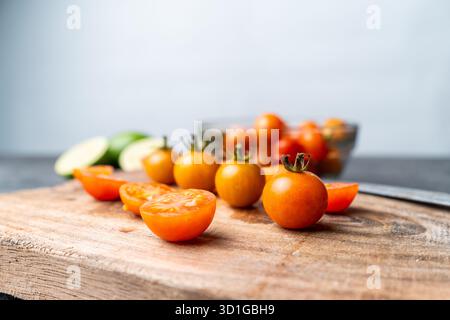 Close-up of fresh cherry tomatoes, whole and sliced, on a rustic wooden cutting board. A knife, bowl, and limes are in the soft bokeh background. Stock Photo