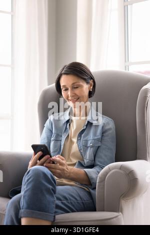 Smiling mature female using smartphone lounging in cozy chair Stock Photo